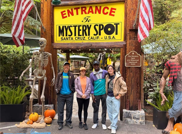 Five people pose for a picture in front of a sign that reads "Entrance To Mystery Spot" with pumpkins and a skeleton.